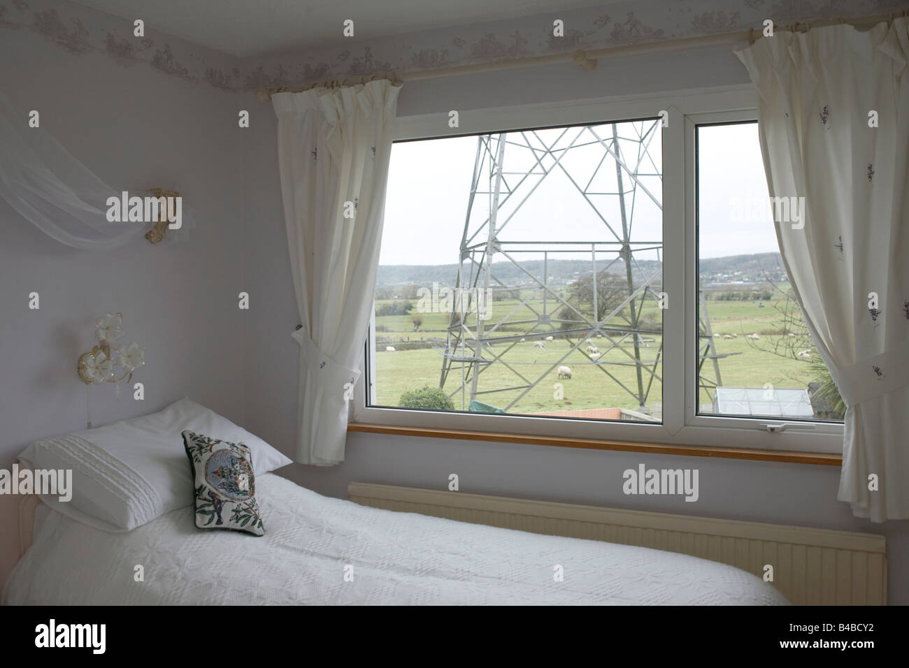 Ein Strommast durch ein Schlafzimmerfenster, wo eine bestickte Kissen lehnt sich an ein Bett, erscheint und über weiden Schafe Stockfoto