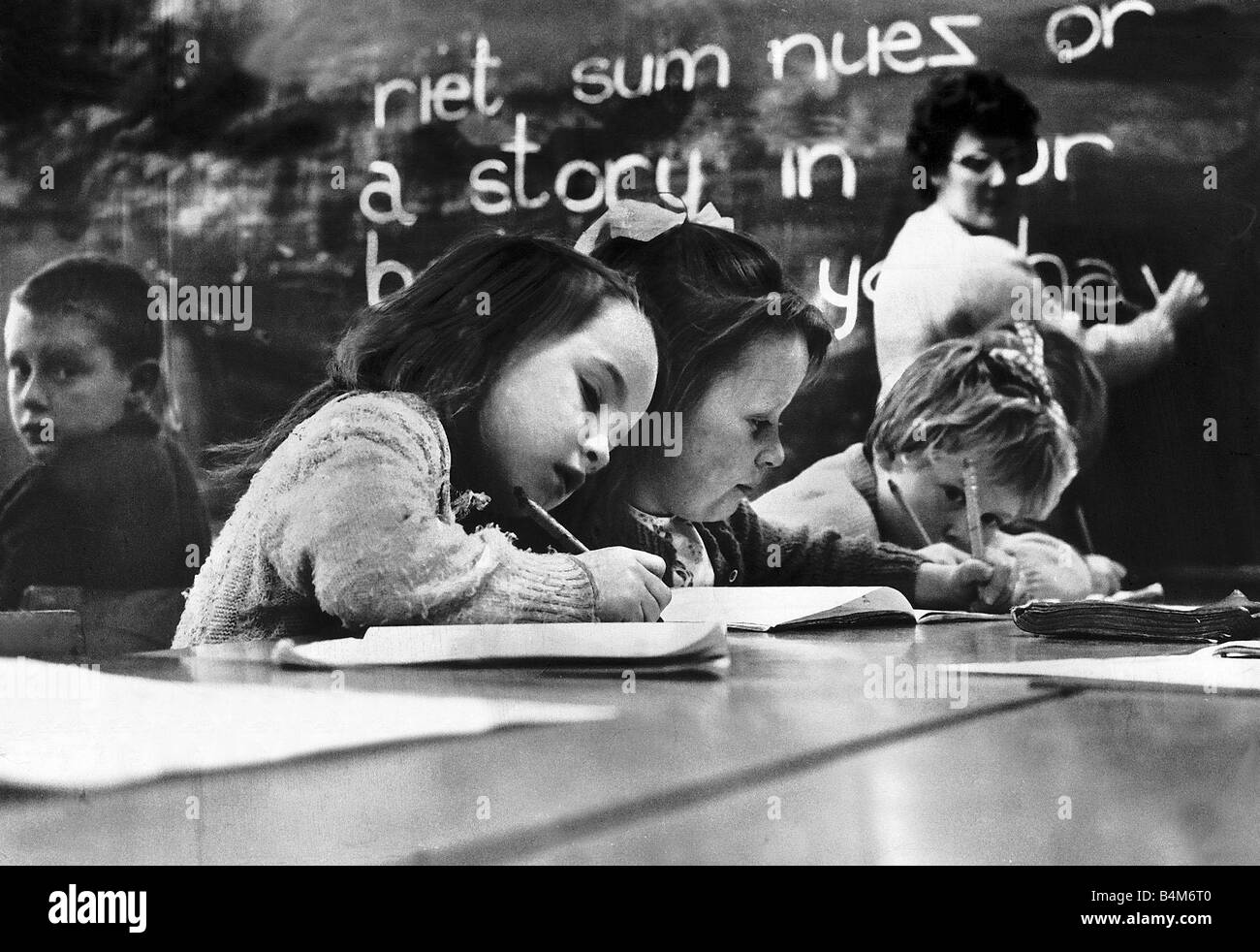 Kinder im Grundschulalter schreiben am Schreibtisch mit Lehrer im Hintergrund auf einer Tafel 1964 schreiben Stockfoto