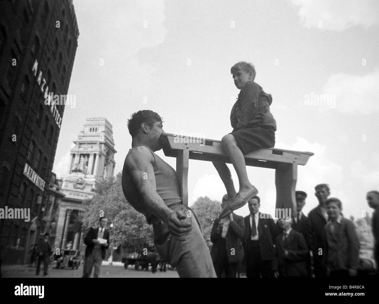 Eine Straße Entertainer am Tower Hill in London hält das Gewicht der ein Junge sitzt auf einer Holzbank mit nur seinen Mund beobachtet von Stockfoto