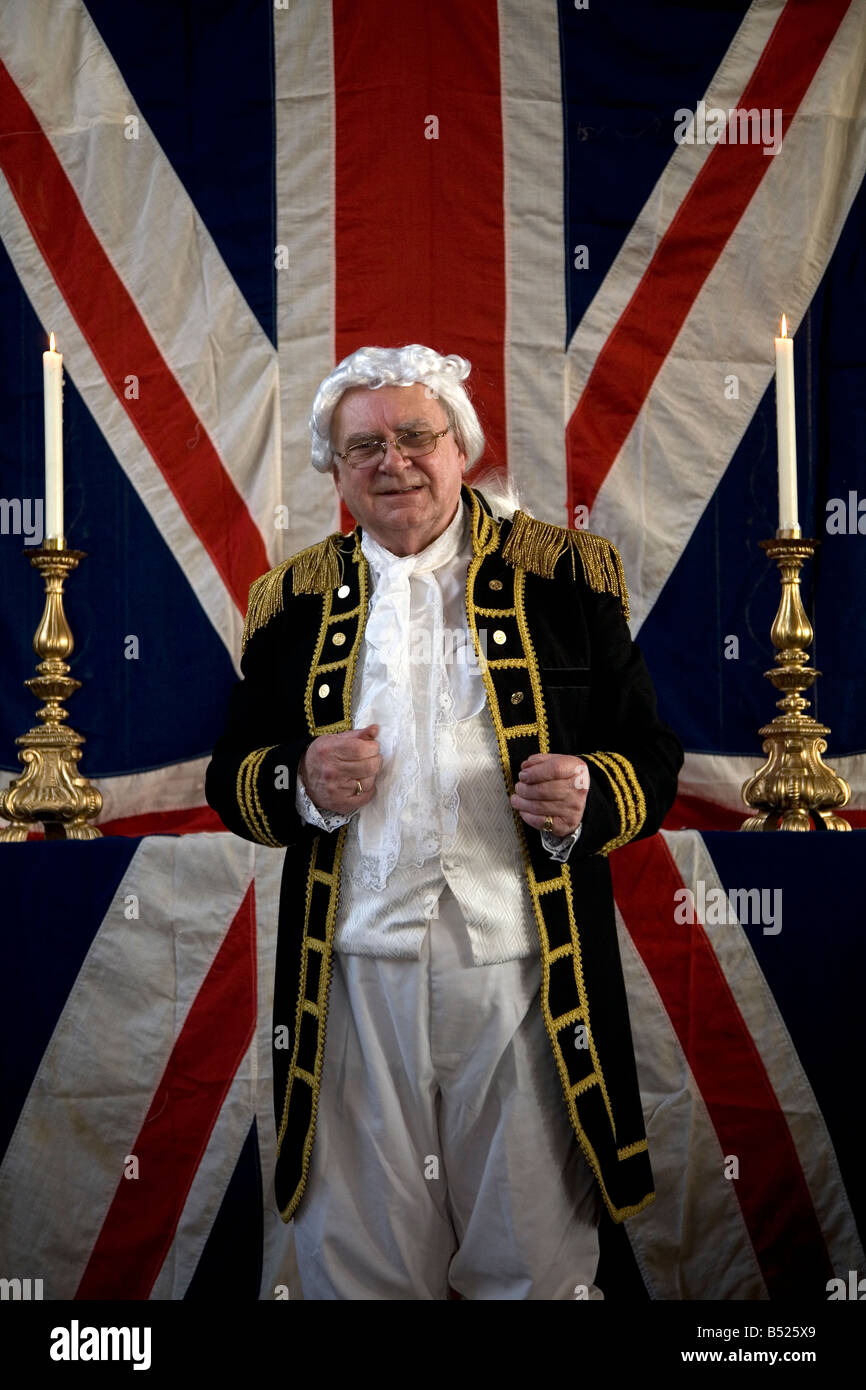 Ein Gentleman gekleidet im Kostüm des a Königliche Marine Offizier steht vor der Nationalflagge am Jahrestag des Trafalgar. Stockfoto