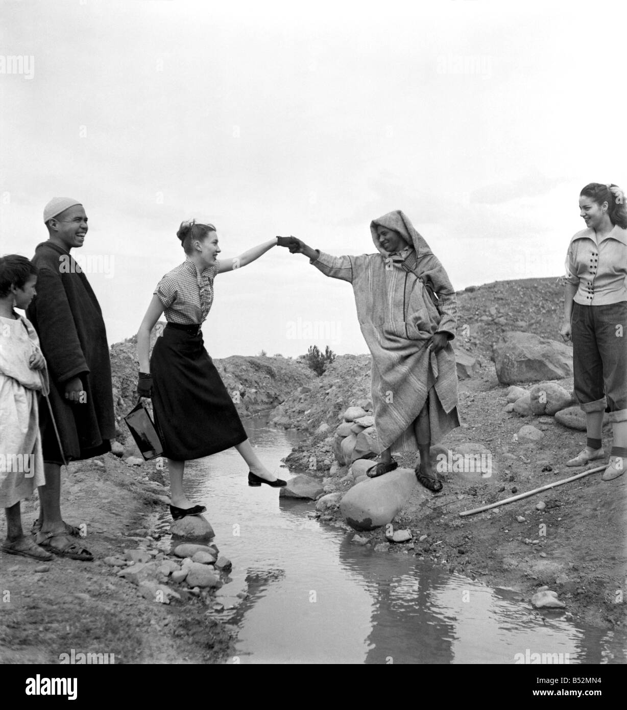 Mann in arabischer Kleidung verleiht seine Hand um französischen Schauspielerinnen über einen Bach zu helfen, als er sie führt auf einer Tour durch die umliegende Landschaft in Marokko.; D Ecember 1952 C5919-050 Stockfoto