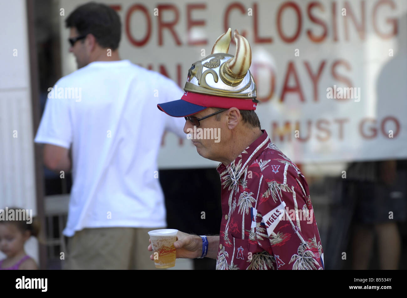 Ein Mann mittleren Alters tragen eine Baseball-Cap mit einem albernen Kunststoff Wikinger Hut mit Hörnern trägt ein voller Bier im Plastikbecher. Stockfoto