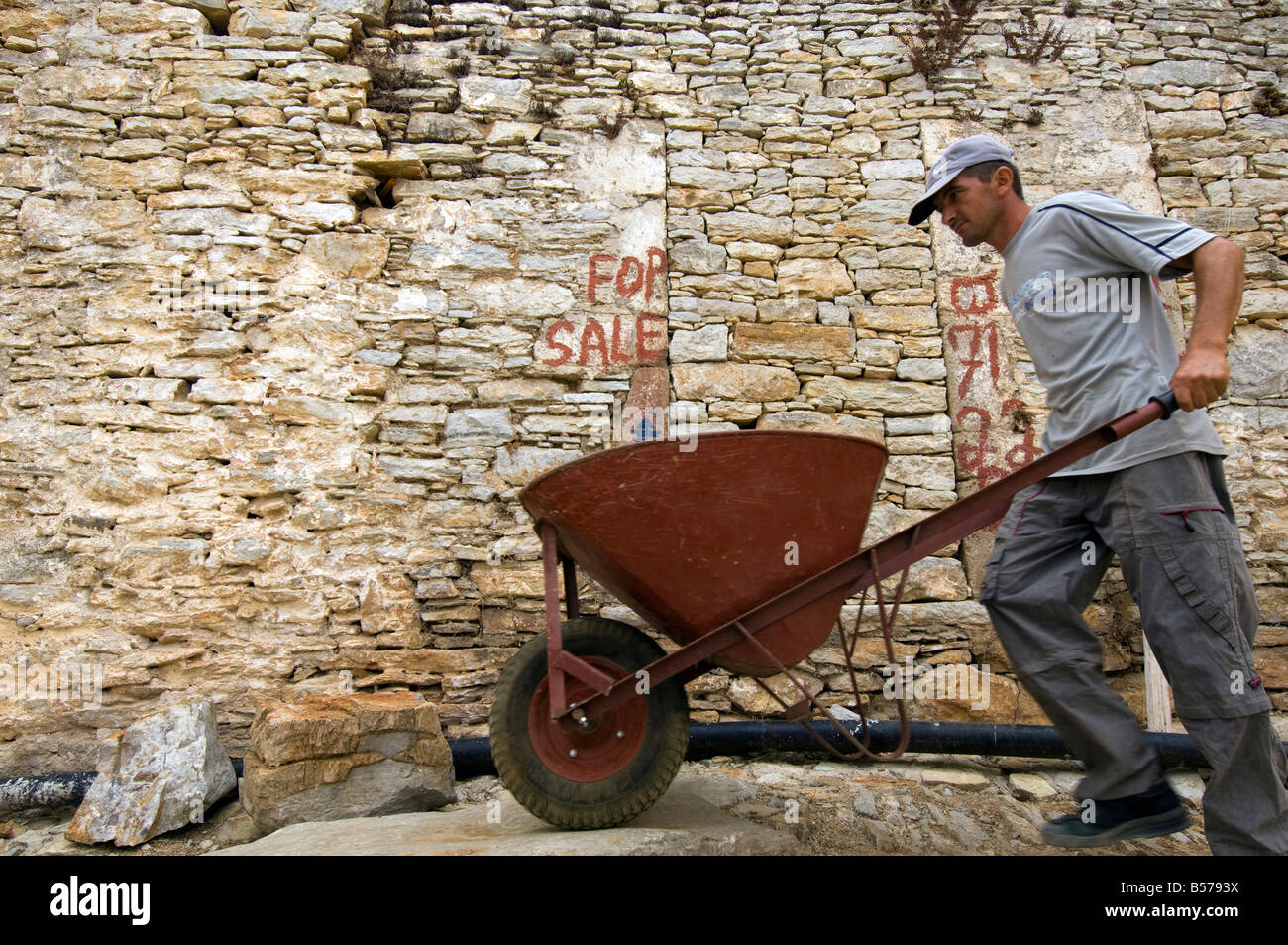 Ein Baumeister schiebt seine Schubkarre vorbei ein steinernes Haus zum Verkauf in die Altstadt oder Chorio, auf der griechischen Insel Symi Stockfoto
