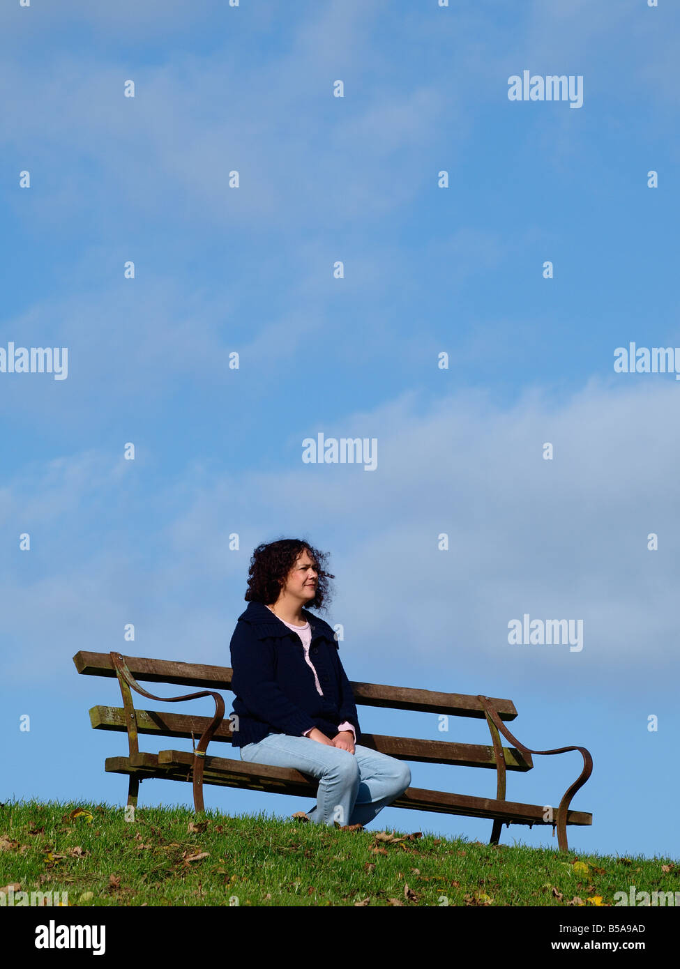 Frau in ihren 30ern sitzt auf einer Parkbank mit einem blauen Himmel gesichert Stockfoto