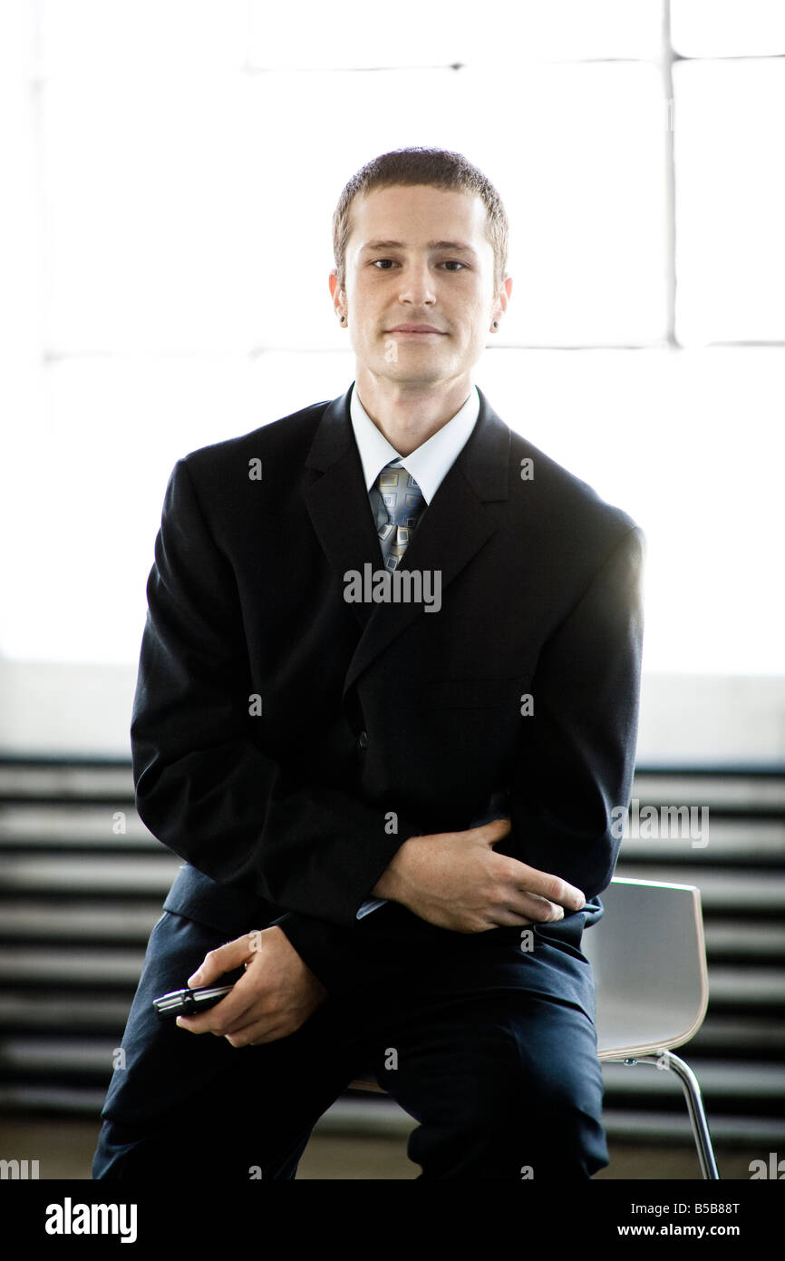 30 Jahre alten Kaukasischen Männchen in einem Business-Anzug sitzt auf Schemel vor großen Fenstern mit einem Mobiltelefon Stockfoto