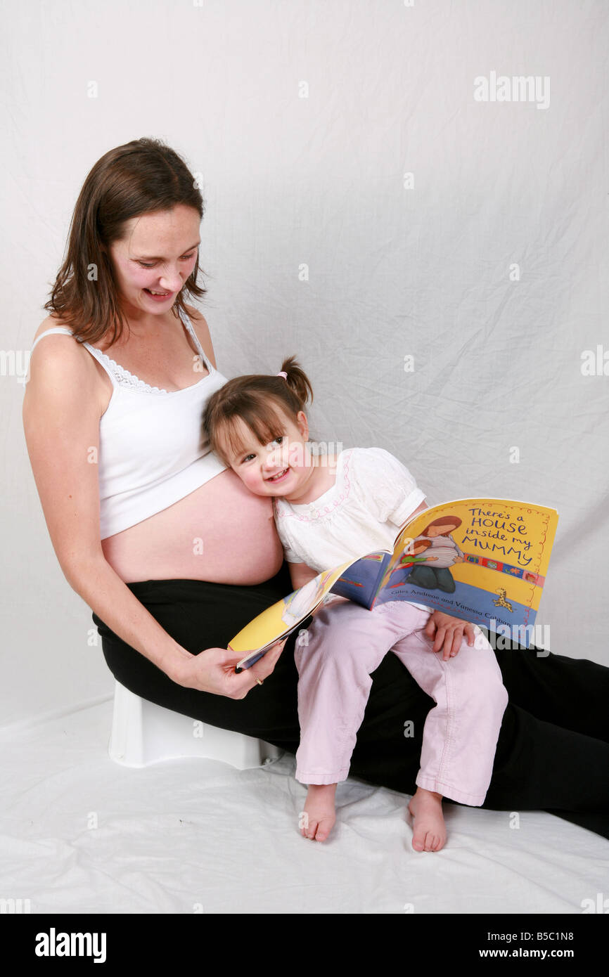 Zwei Jahre sitzt alte Tochter Kleinkind auf schwangere Mütter Schoß Kinderbuch auf Schwangerschaft Magen Fötus Geräusche hören lesen Stockfoto