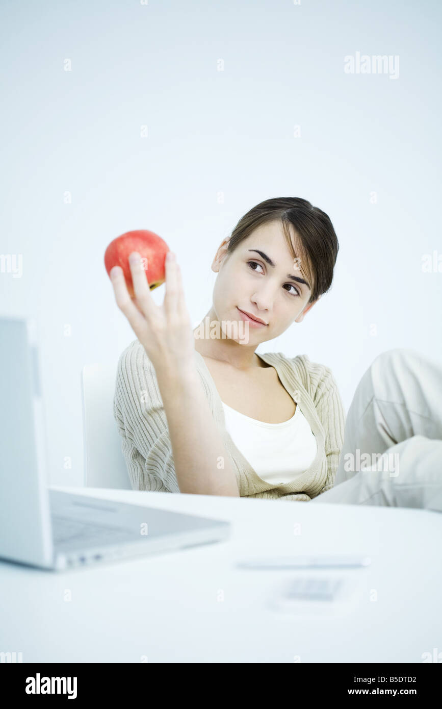 Junge Frau sitzt am Schreibtisch, mit Blick auf Apfel in der hand Stockfoto