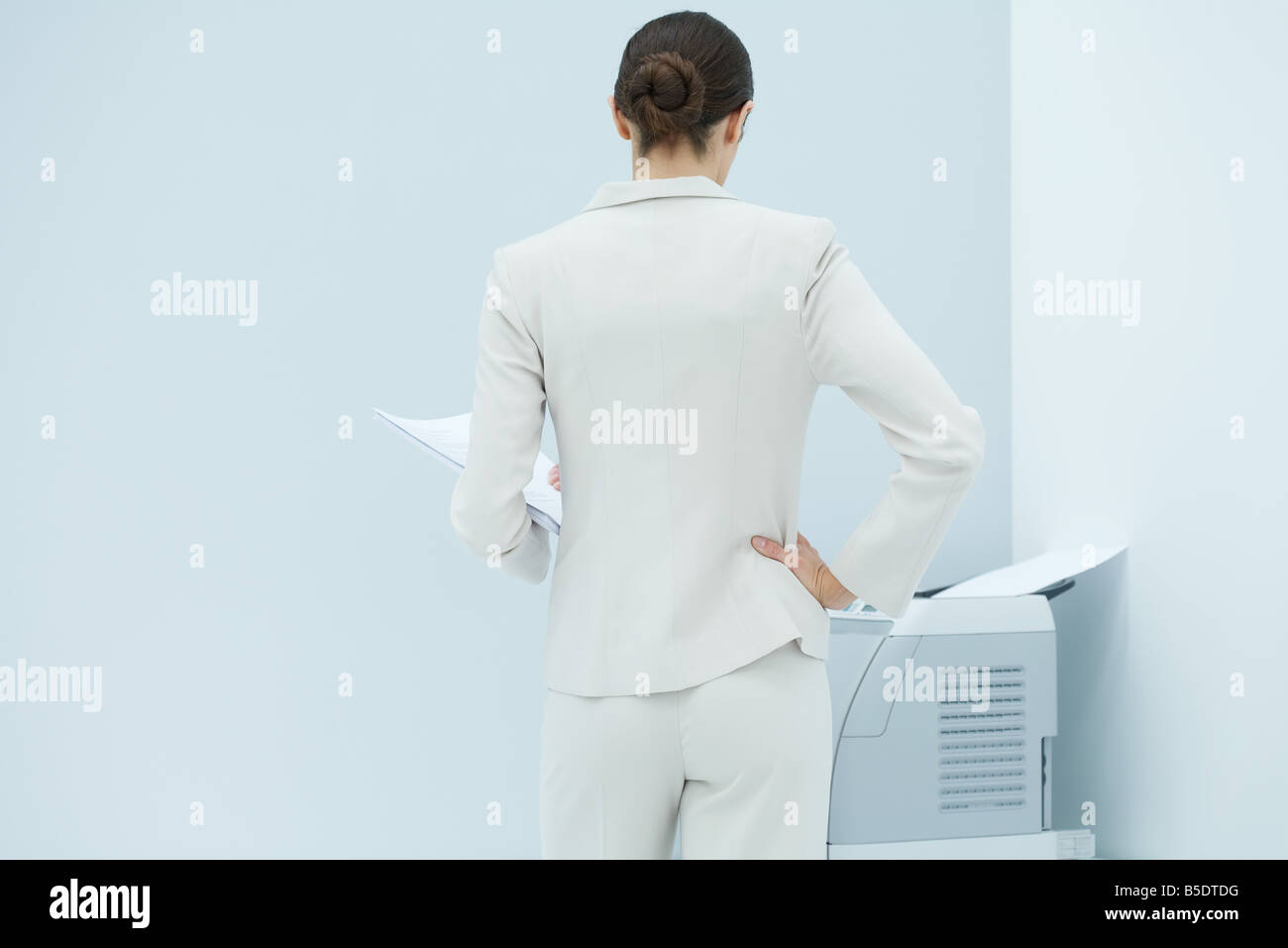 Geschäftsfrau mit Hand auf Hüfte stehend, mit Blick auf Drucker, Rückansicht Stockfoto