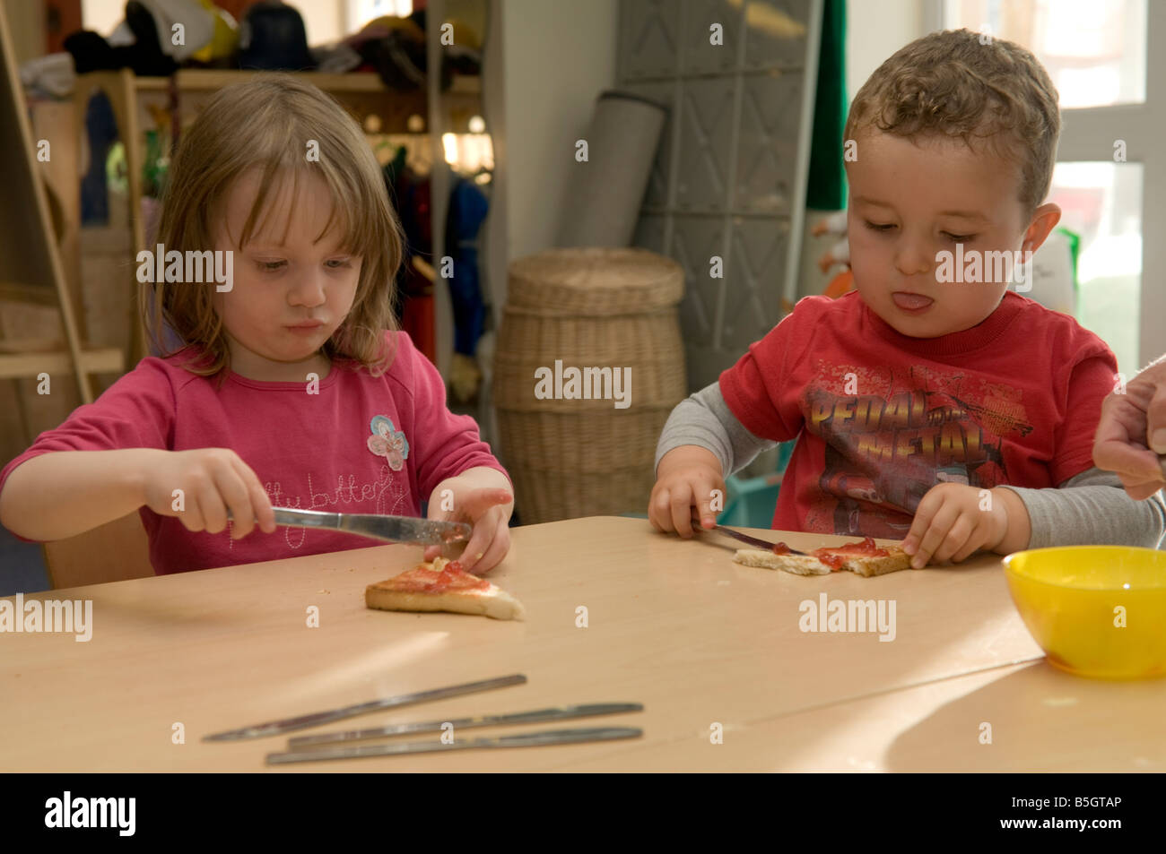 Zwei 2 kleinen Kindern im Kindergarten Kindertagesstätte verbreiten Marmelade auf ihre Morgen Toast zum Frühstück, erlernen grundlegende Fähigkeiten, UK Stockfoto