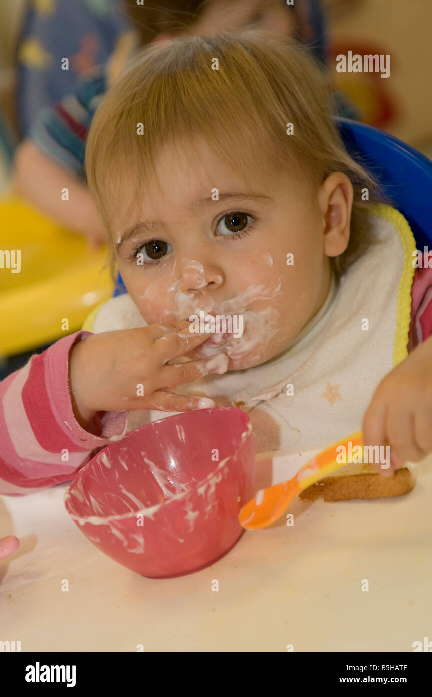 jungen Baby Kleinkind Kind im Kindergarten Kita Zentrum Kleinkind Essen Joghurt messily tragen Lätzchen, chaotisch Gesicht, UK Stockfoto