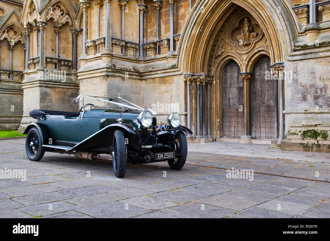 Ein renovierter, flaschengrüner Lagonda-Oldtimer aus den 1930er Jahren, der mit weißen Hochzeitsbändern vor der Wells Cathedral in Somerset, England dekoriert ist. Stockfoto