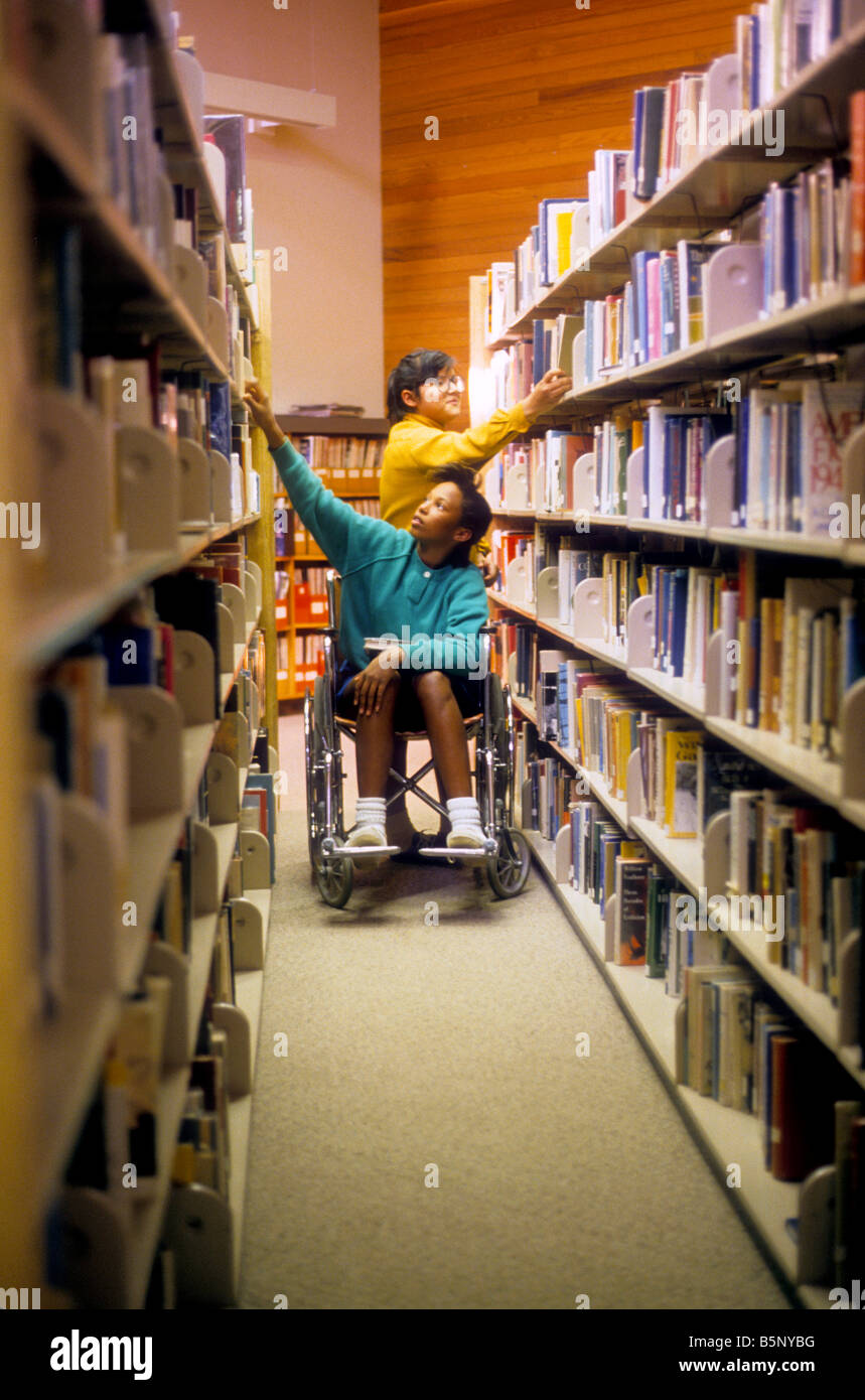 Schwarze Mädchen im Rollstuhl und hispanischen Mädchen wählen Sie Bücher aus den Regalen der Schulbibliothek. Stockfoto