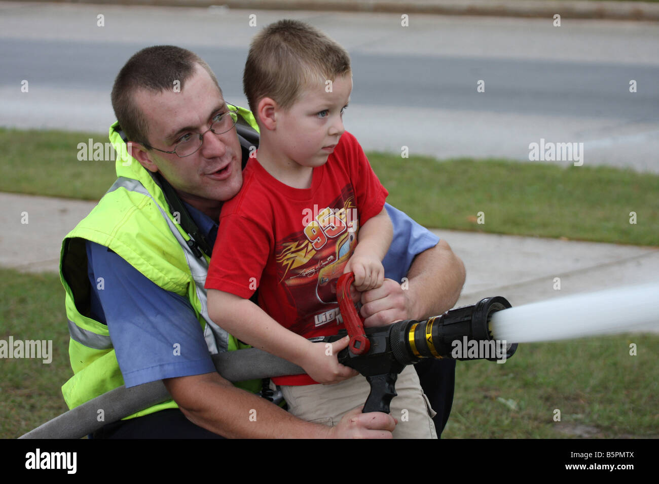 Ein Feuerwehrmann hilft einen kleiner Junge der Feuerwehrschlauch zu verwenden, um eine Demonstration Feuer auf einem Feuer Sicherheit Messe Stockfoto