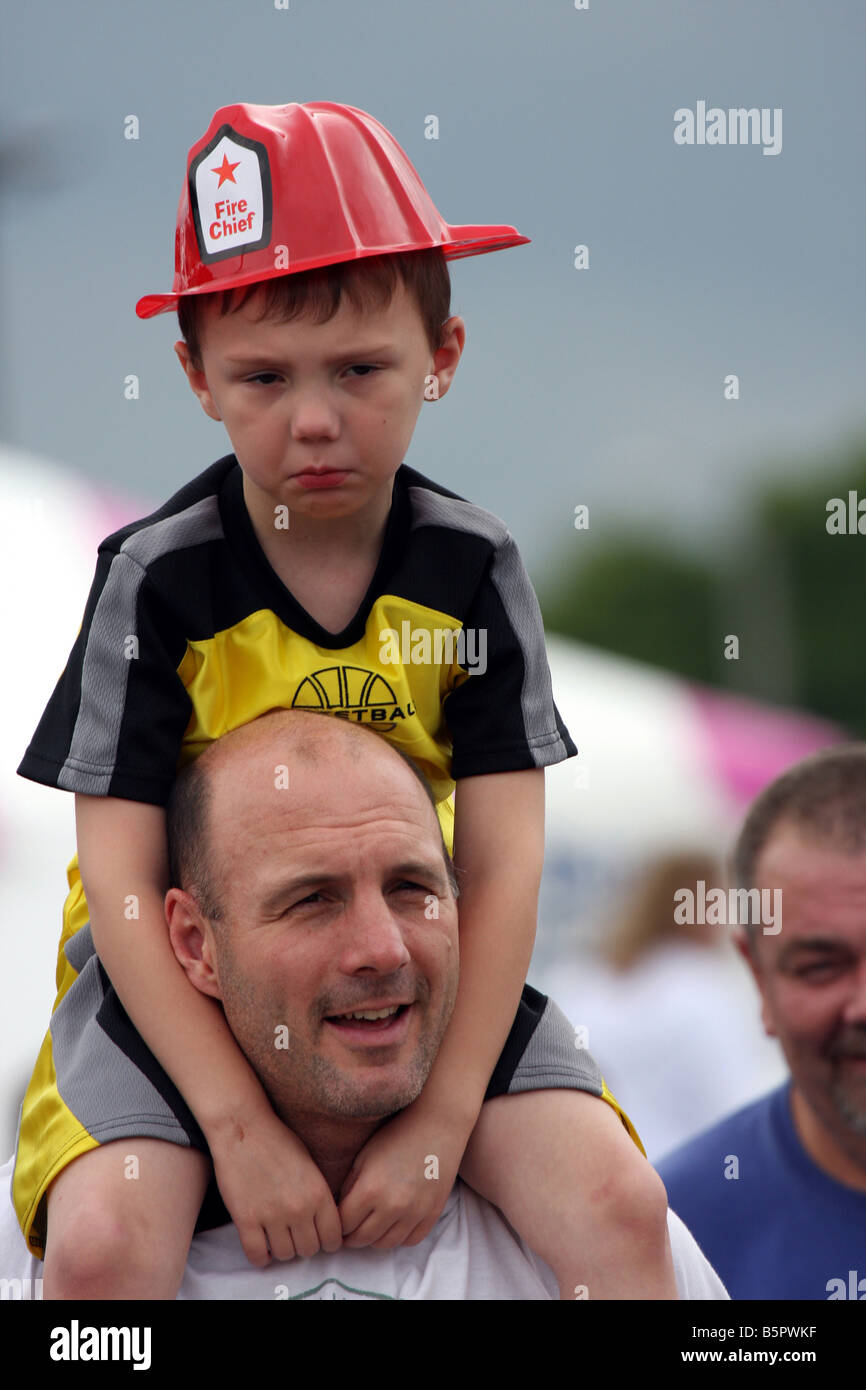 Ein kleiner Junge auf den Schultern von seinem Vater wütend oder traurig über etwas auf ein Feuer Sicherheit Messe suchen Stockfoto
