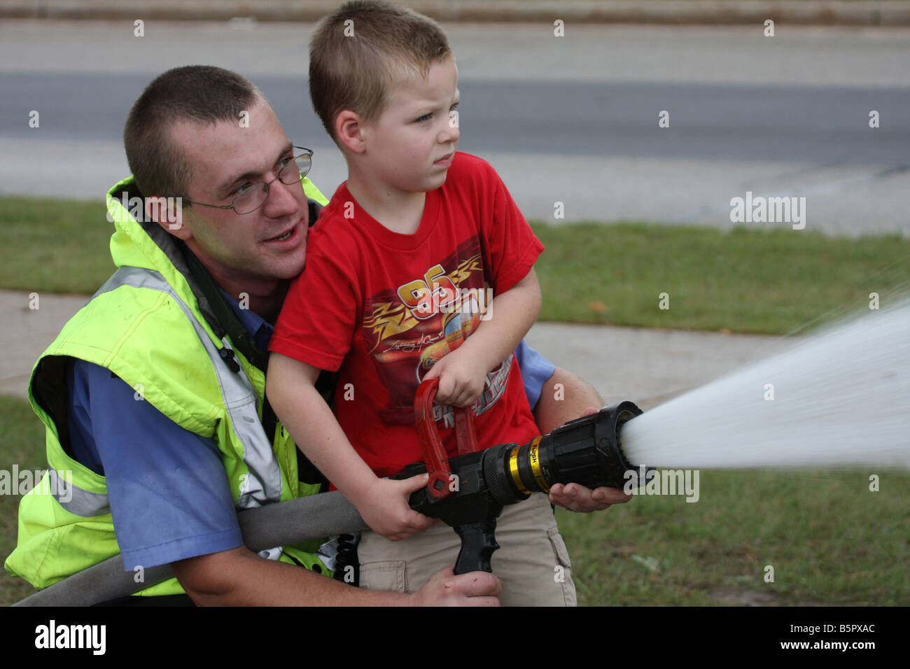 Ein Feuerwehrmann hilft einen kleiner Junge der Feuerwehrschlauch mit solcher Entschiedenheit zu verwenden, um löschte eine Demonstration Feuer bei einem Feuer-Sicherheit-Fai Stockfoto