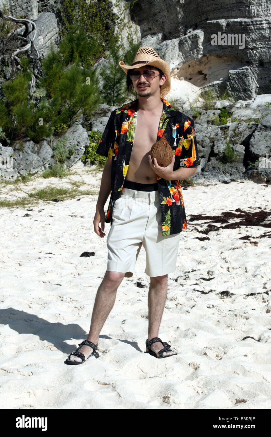 Junger Mann trug Bermuda-Shorts, ein Hawaii-Hemd und einen Strohhut hält eine Kokosnuss am Strand von Stonehole, Bermuda Stockfoto