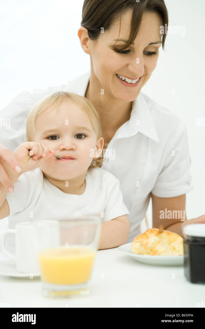 Berufstätige Frau sitzt am Frühstückstisch, Kleinkind auf Schoß, Löffel im Mund Stockfoto
