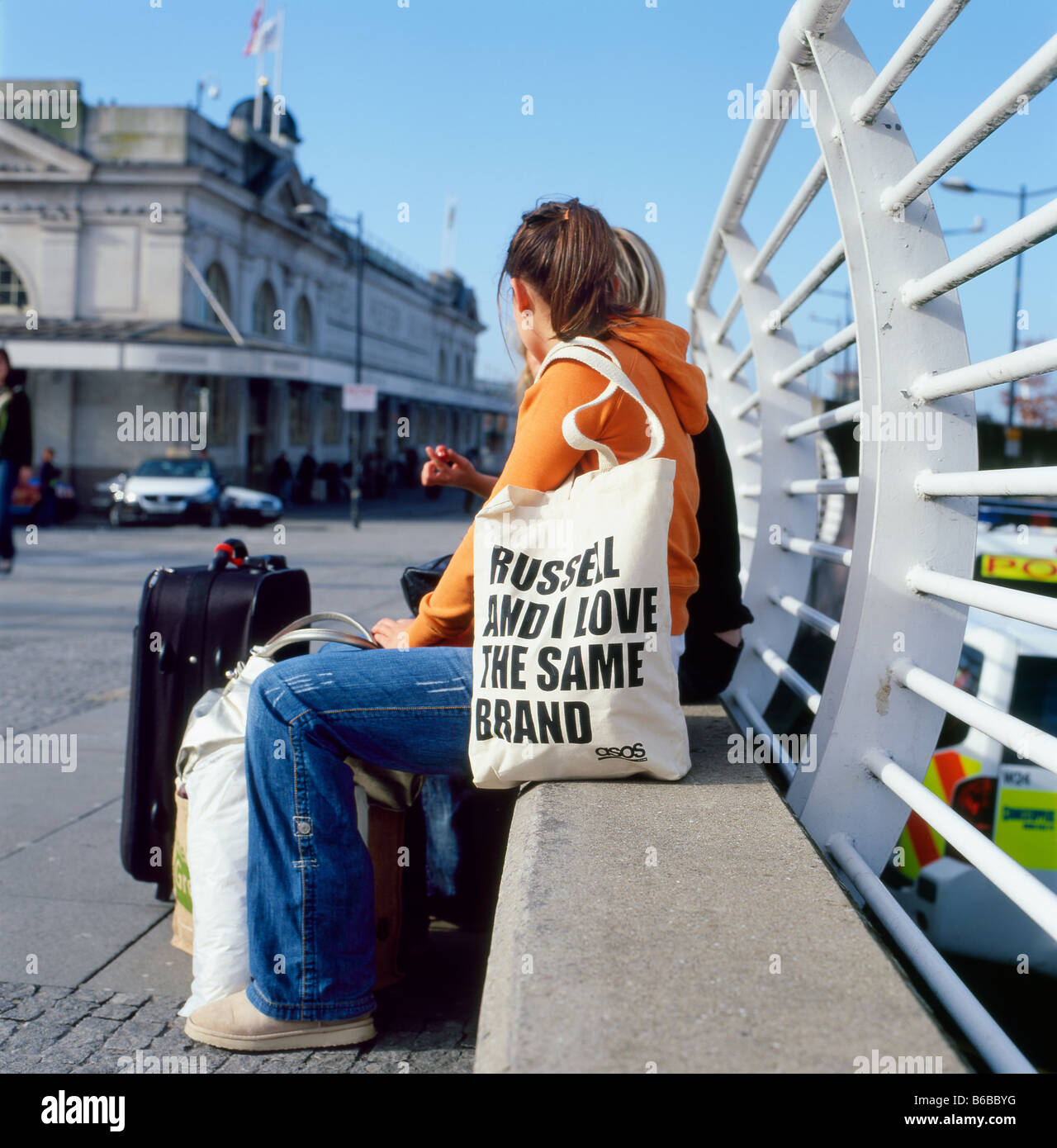 Eine junge Frau, die an einer Wand in der Nähe des Bahnhofs sitzt und eine Einkaufstasche der Marke Russell in Cardiff Großbritannien, Großbritannien, KATHY DEWITT, trägt Stockfoto