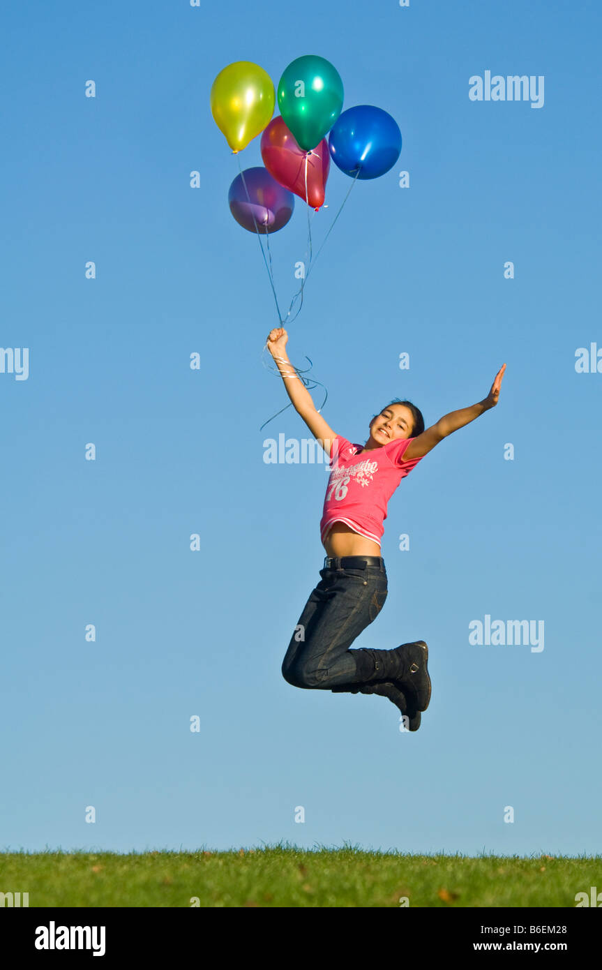Ein junges Mädchen (11) springen in der Luft mit einer Reihe von bunten Helium gefüllte Ballons vor blauem Himmel. Stockfoto