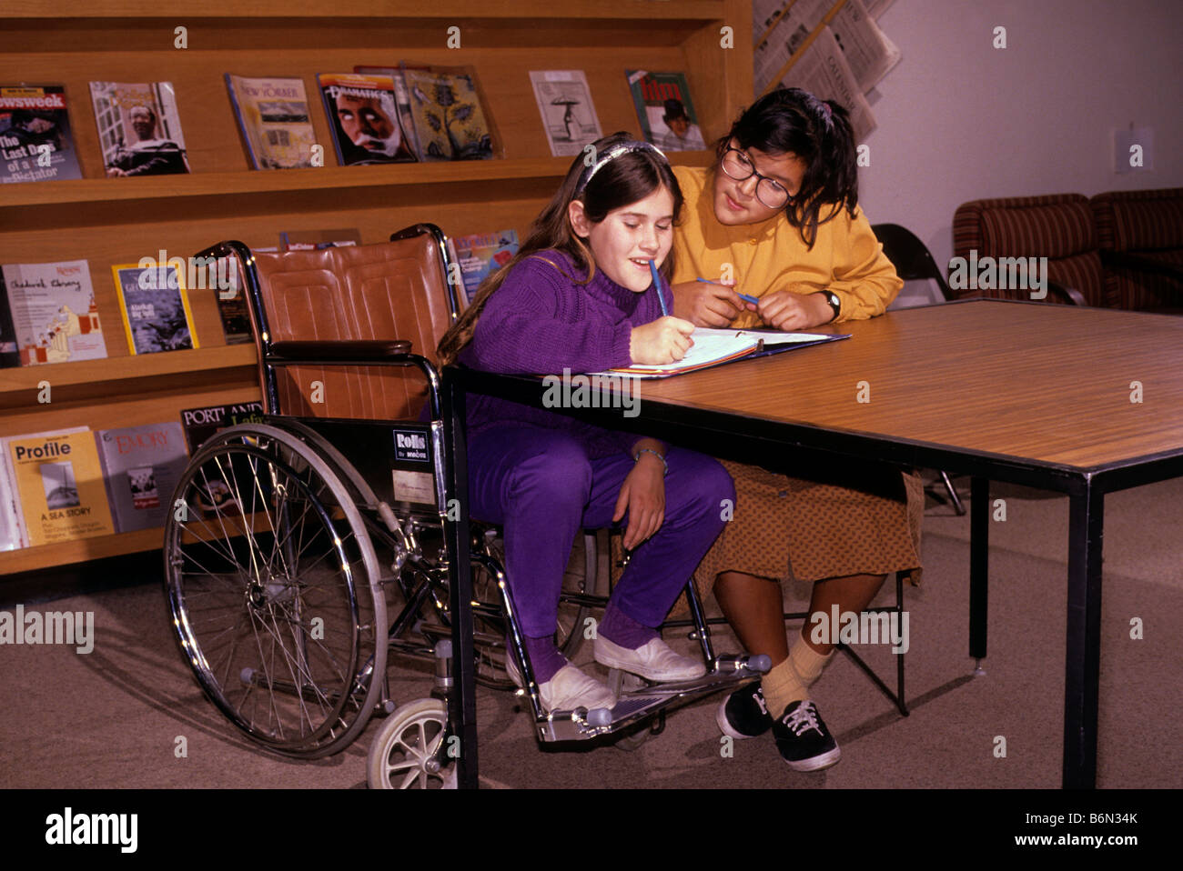 Hispanischen Mädchen hilft weißes Mädchen im Rollstuhl, die Schule zu tun Arbeit in der Schulbibliothek Stockfoto