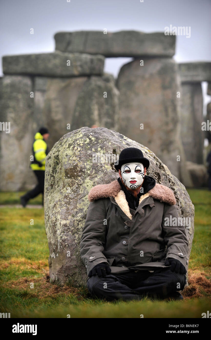 EIN BESUCHER NACH STONEHENGE MIT WHITEFACE CLOWNSMASKE SITZT DURCH EINE ÄUßERE STATION STEIN, WÄHREND EIN WACHMANN SCHEINT BEKANNTGEBEN Stockfoto