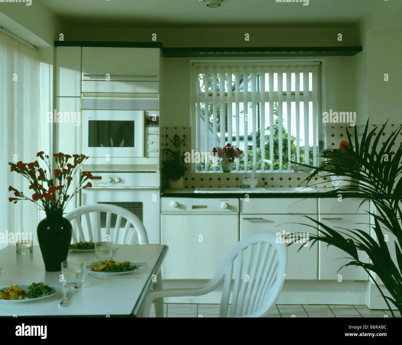 Weiße Stühle und Tisch in weiße Küche mit Lattenrost Rollo am Fenster oben ausgestattet Geschirrspüler Stockfoto
