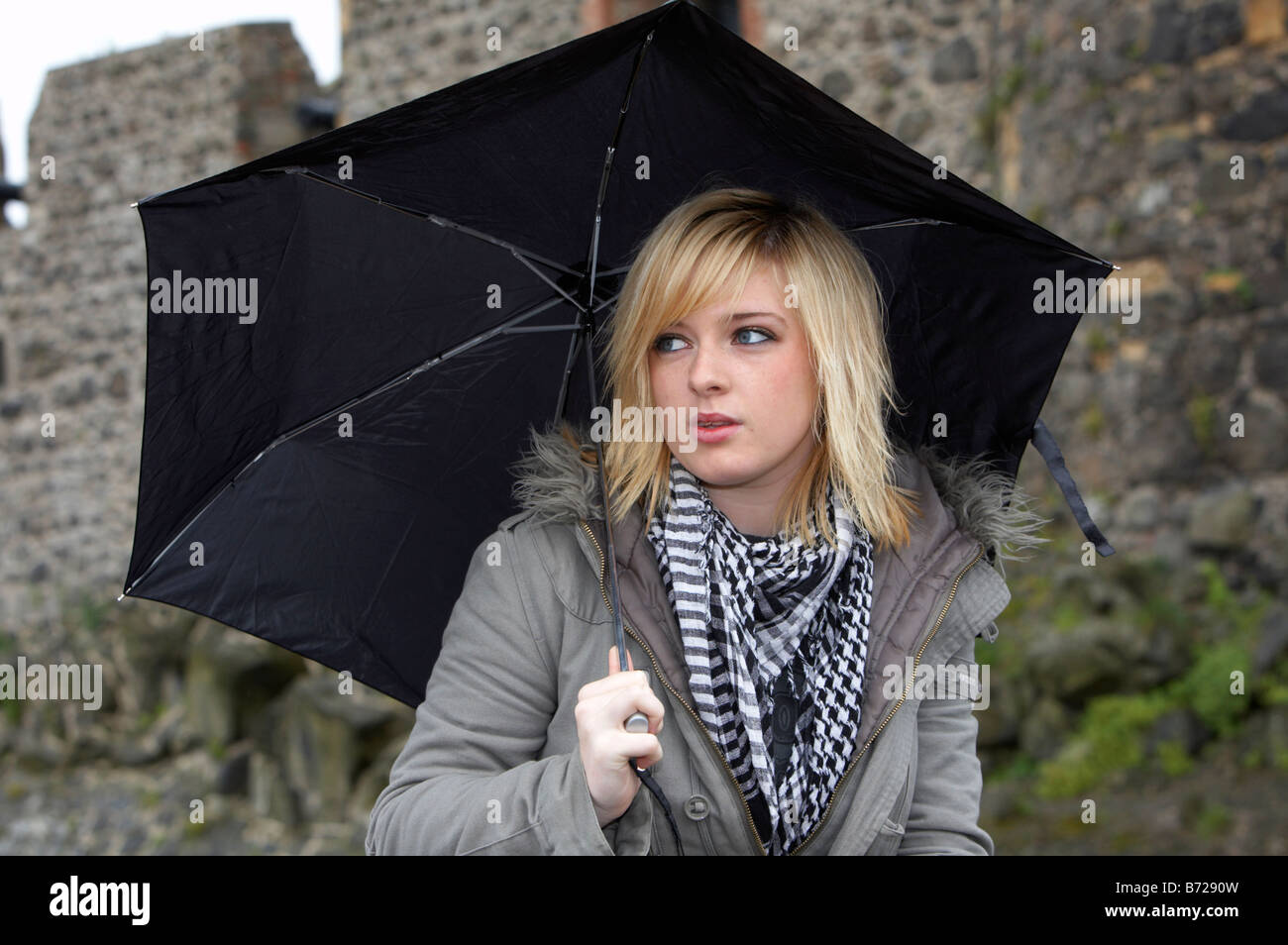 18 jährige Blondine hält Regenschirm tragen Jacke und schwere Schal wegsehen von Kamera in der Frontseite Carrickfergus castle Stockfoto