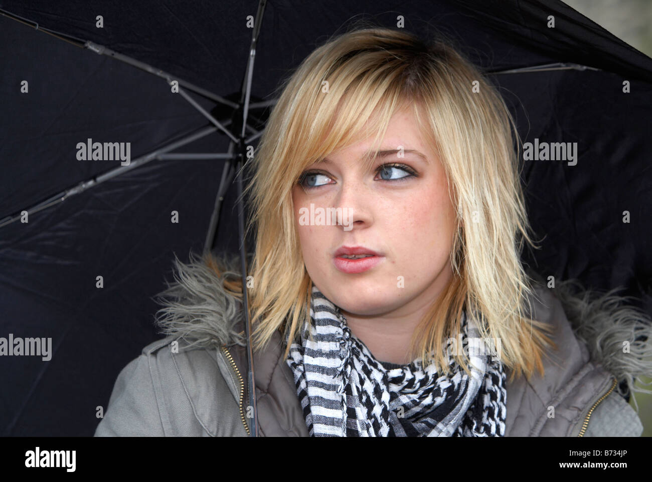 18 jährige Blondine mit schwarzen Regenschirm tragen Jacke und schwere Schal wegsehen von Kamera Stockfoto