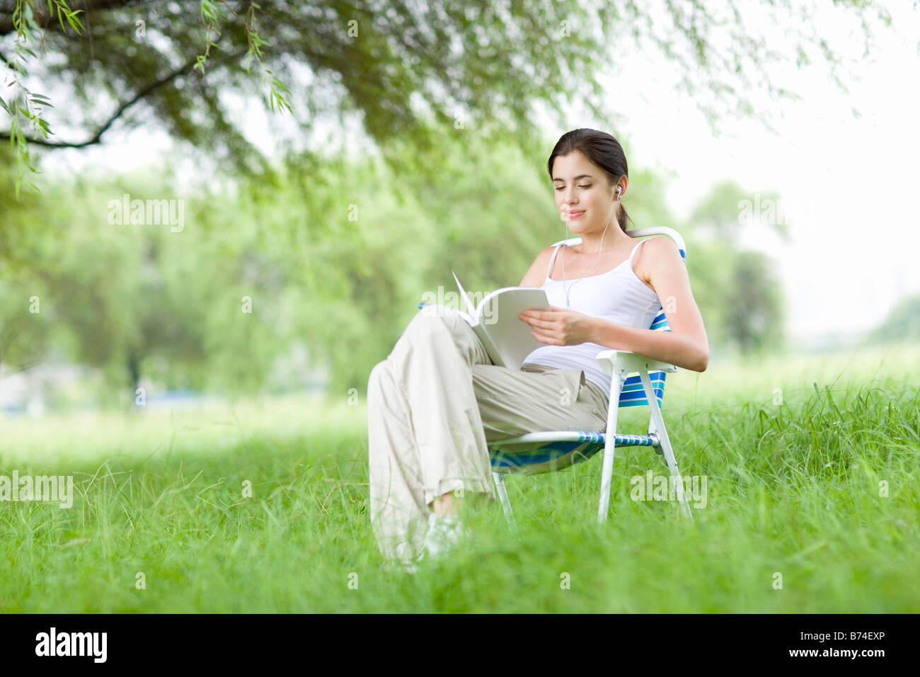 Junge Frau sitzt auf Liegestuhl hören mp3-Player mit Buch im freien Stockfoto