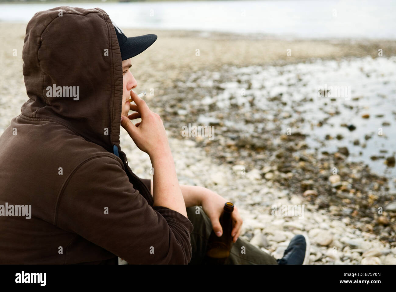 einsame junge sitzt am Ufer und Bier aus einer Flasche trinkt und raucht eine Zigarette Stockfoto