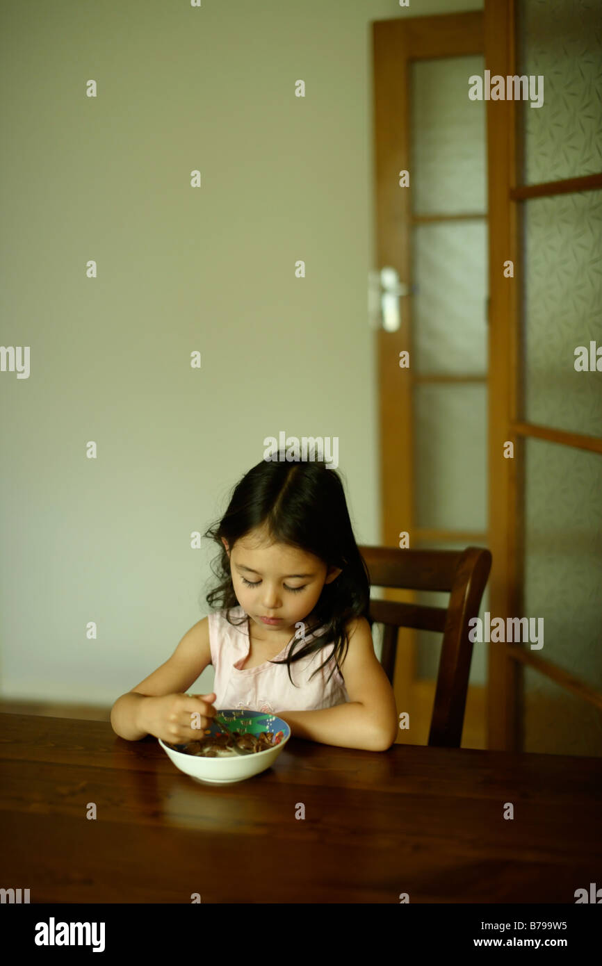 Fünf Jahre altes Mädchen sitzt auf einem Holztisch und isst eine Schüssel Müsli Stockfoto