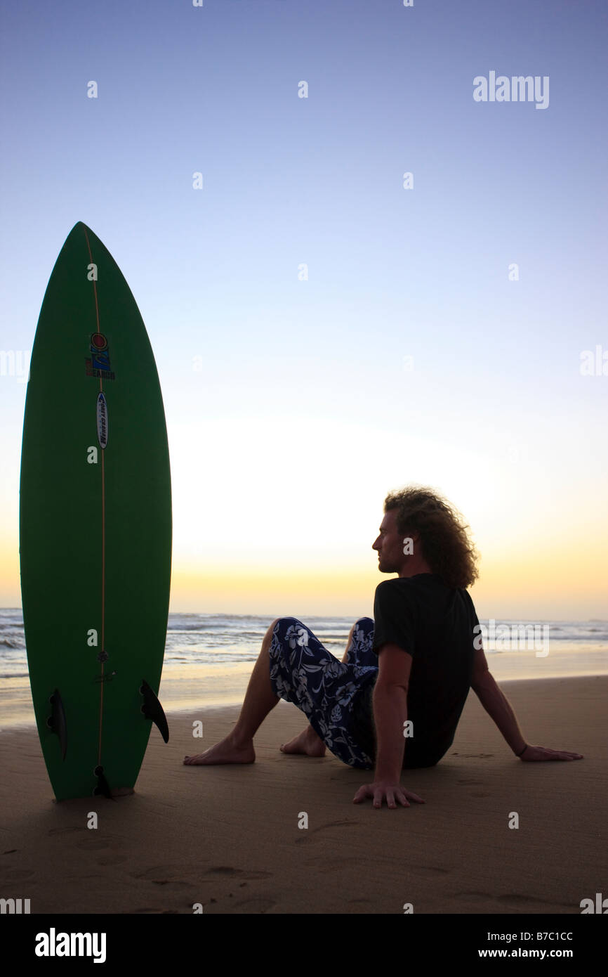 ein junger Mann mit seinem Surfbrett den Sonnenuntergang am Strand sitzen. Stockfoto