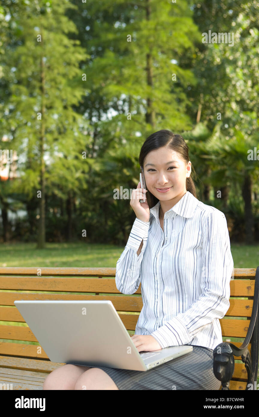 Eine Business-Frau sitzt auf der Bank mit Mobiltelefon und Laptop in die Kamera schaut Stockfoto