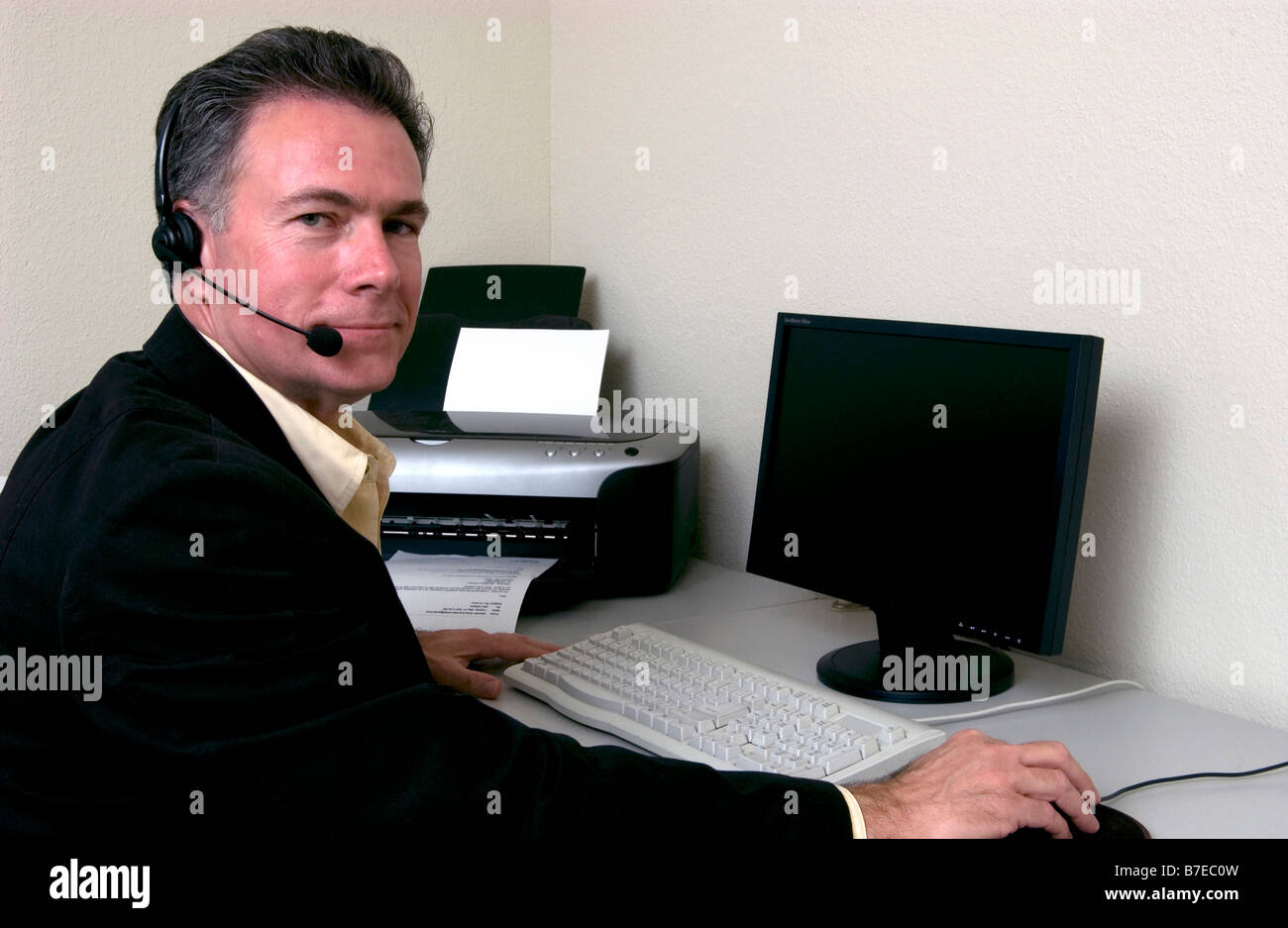 Ein Mann sitzt an einem Schreibtisch vor dem Computer mit einem Kopfhörer auf und einem freundlichen Lächeln Stockfoto