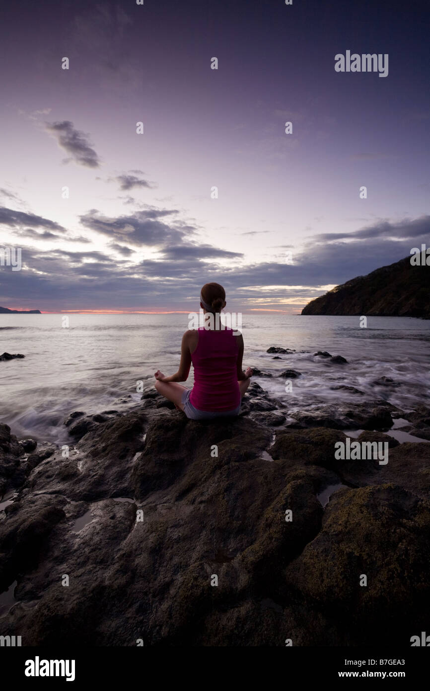 Junge Frau, meditieren auf Felsen in der Abenddämmerung vor der Meeresbrandung in Playas del Coco, Costa Rica. Stockfoto