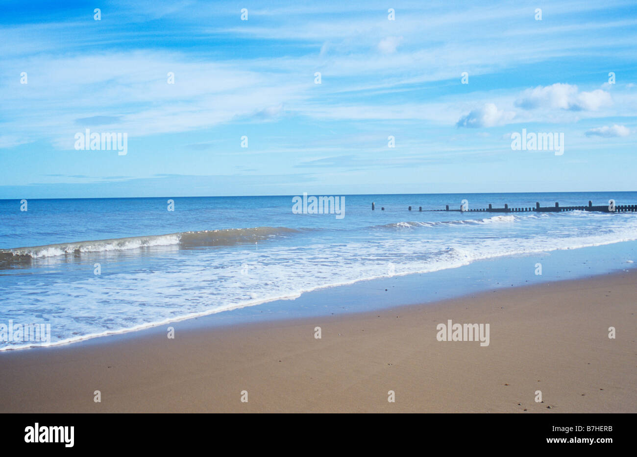 Sanft plätschernde Wellen plätschern auf saubere menschenleeren Sandstrand mit Buhne unter blassen blauen Himmel mit Wolken gestreift Stockfoto