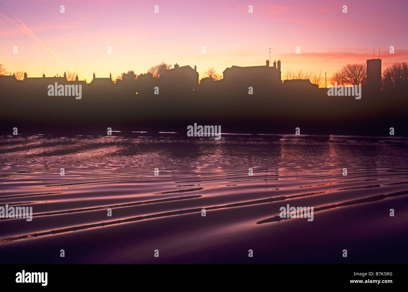 Magenta Sonnenuntergang Himmel spiegelt sich in plätschernden Fluss mit Streifen der Stadtentwicklung in-between Silhouette Stockfoto