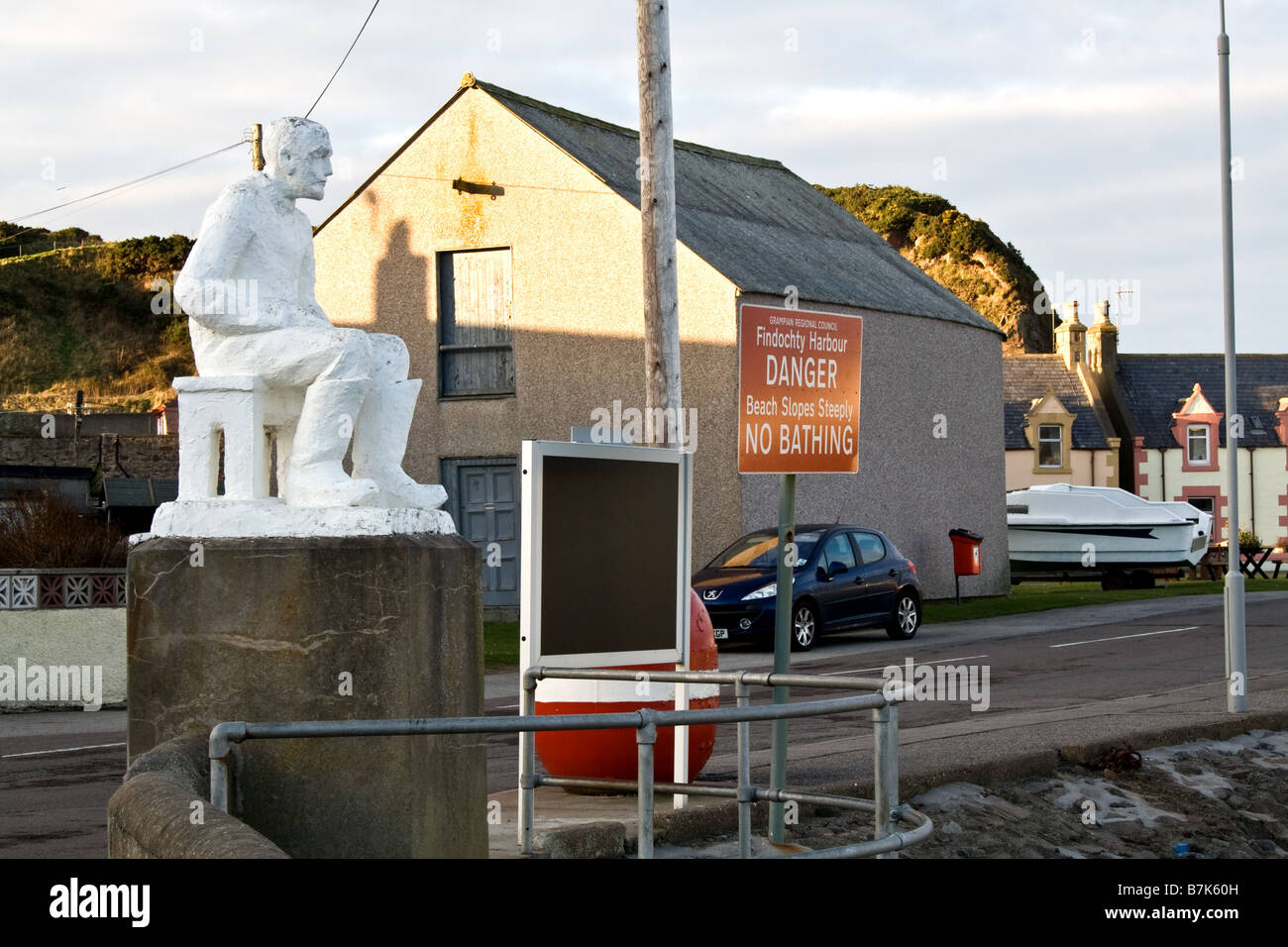 Statue eines Mannes sitzen auf einer Bank neben einem roten Warnschild Blick in Findochty, Schottland Stockfoto