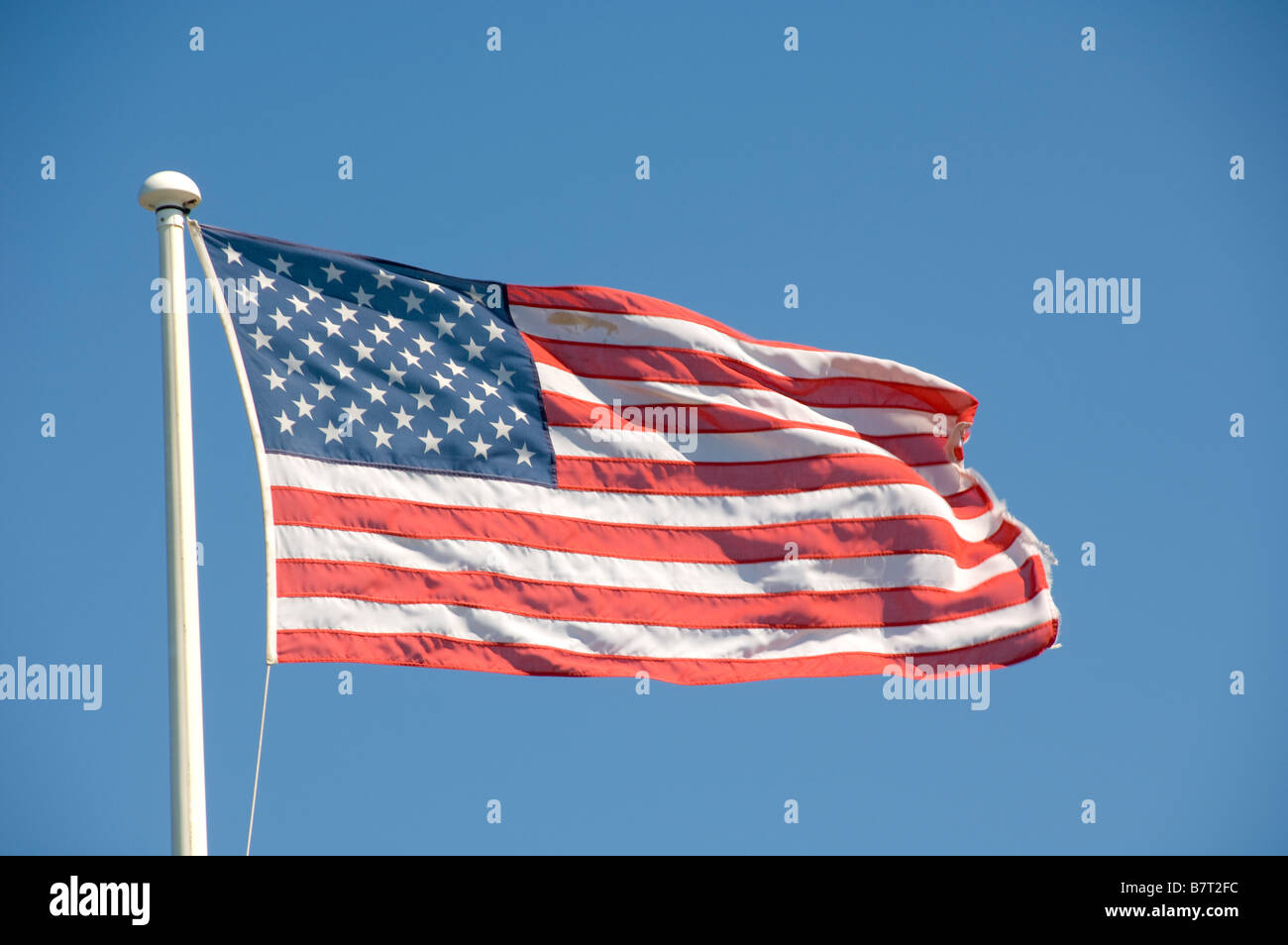 Verblasste amerikanische Flagge, die auf einem weißen Fahnenmast vor blauem Himmel fliegt. Stockfoto