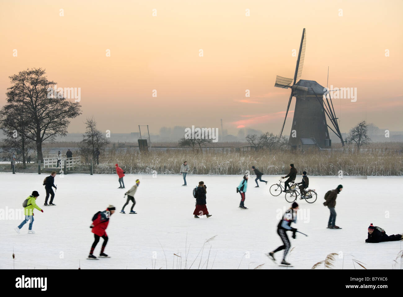 Eisläufer und auch Radfahrer auf Natureis vor einer Windmühle bei Kinderdijk Holland, Niederlande. Bei Sonnenuntergang. Stockfoto