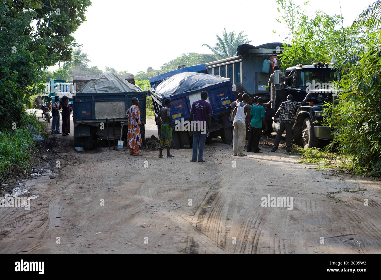 Zwei LKWs haben in einem schlammigen Graben auf der Straße und ein Drittel festgefahren, die LKW quetscht Vergangenheit herunterdrücken Urwaldbäume in Nigeria Stockfoto