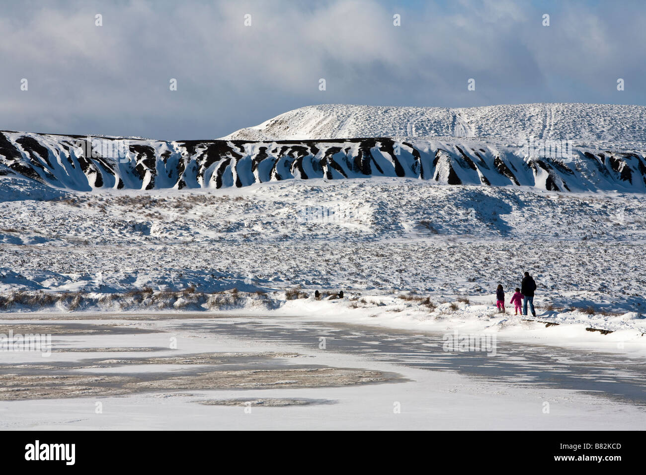 Mann mit zwei Kindern zu Fuß neben zugefrorenen See im Winter mit alten Kohle verwöhnen Tipp hinter Blorenge Wales UK Stockfoto
