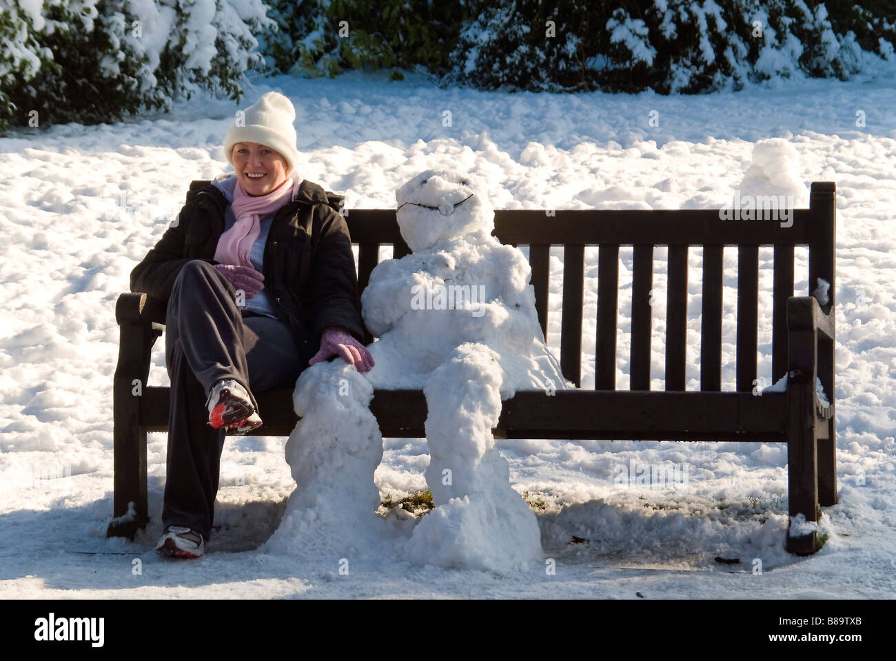 Humorvolle Horizontal Nahaufnahme Portrait eines traditionellen Schneemann und eine junge Frau sitzt auf einer Parkbank in der Sonne Stockfoto