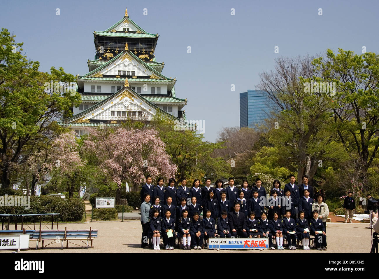 Gruppe japanischer Schulkinder in schwarzer Uniform, die vor dem Schloss Osaka und Kirschblütenbäumen für ein formelles Bild posieren. Frühling. Stockfoto