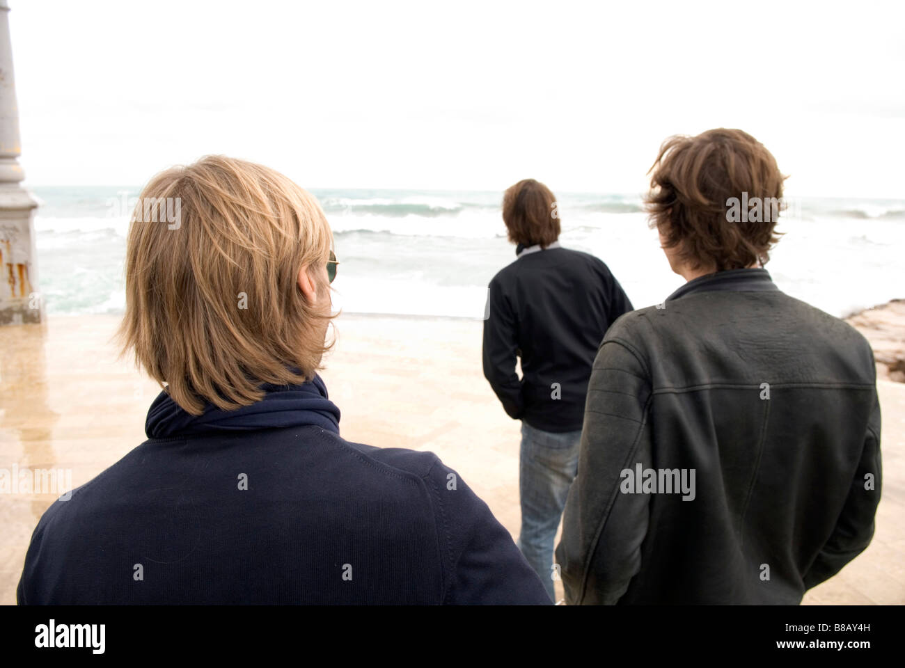 eine Rückansicht von drei erwachsenen Männern stehend auf einer Strandpromenade mit Blick auf das Meer an einem stürmischen Tag Stockfoto