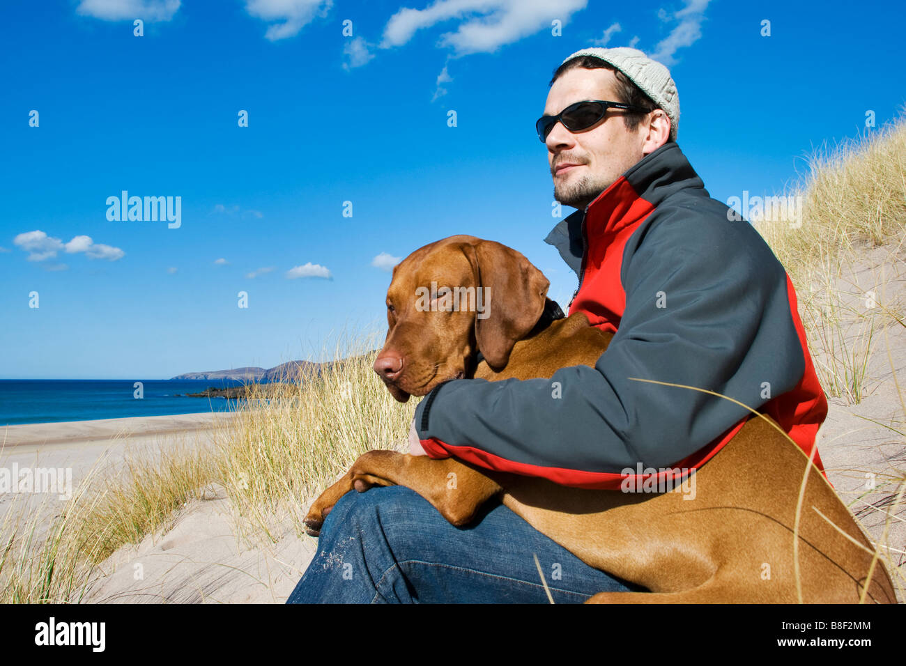 Mann saß mit Magyar Vizsla Hund auf seinem Schoß auf die isolierte uk Strand Sandwood Bay in Schottland Stockfoto