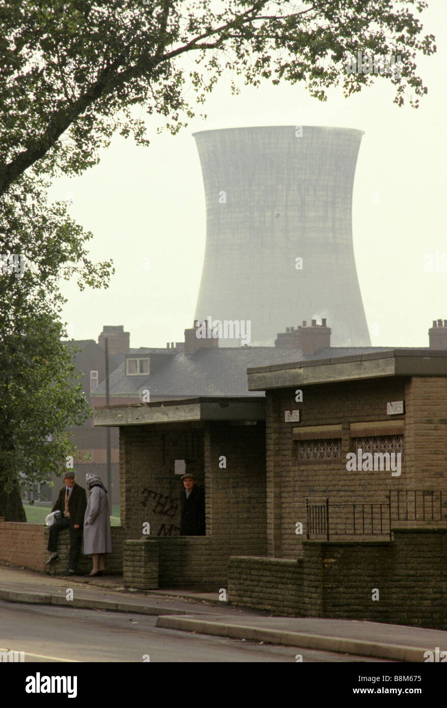 Menschen warten an einer Bushaltestelle in Grimethorpe mit großen Fabrikschornsteinen erhebt sich hinter ihnen. Stockfoto