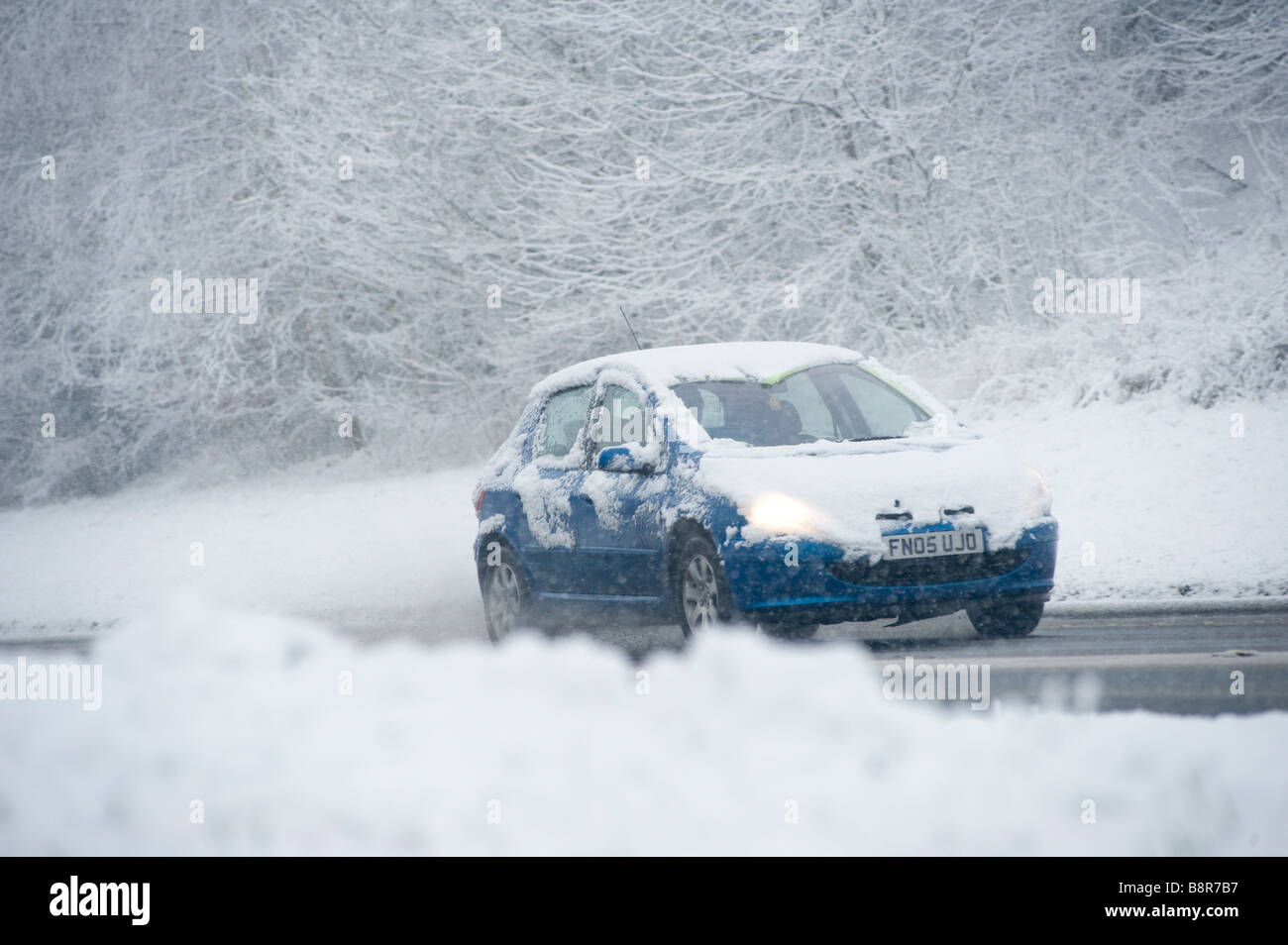 Blaues Auto Fahrt langsam auf einer verschneiten Straße im Winter in England mit Scheinwerfern mit Schnee bedeckt Stockfoto