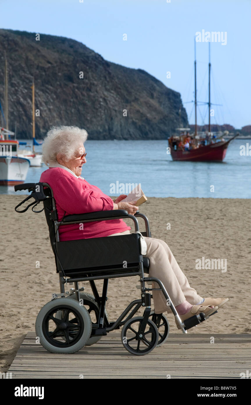 Zufrieden ältere Behinderte Dame sitzt im Rollstuhl ein Buch an einem sonnigen Urlaub Urlaub Strand Meer Stockfoto