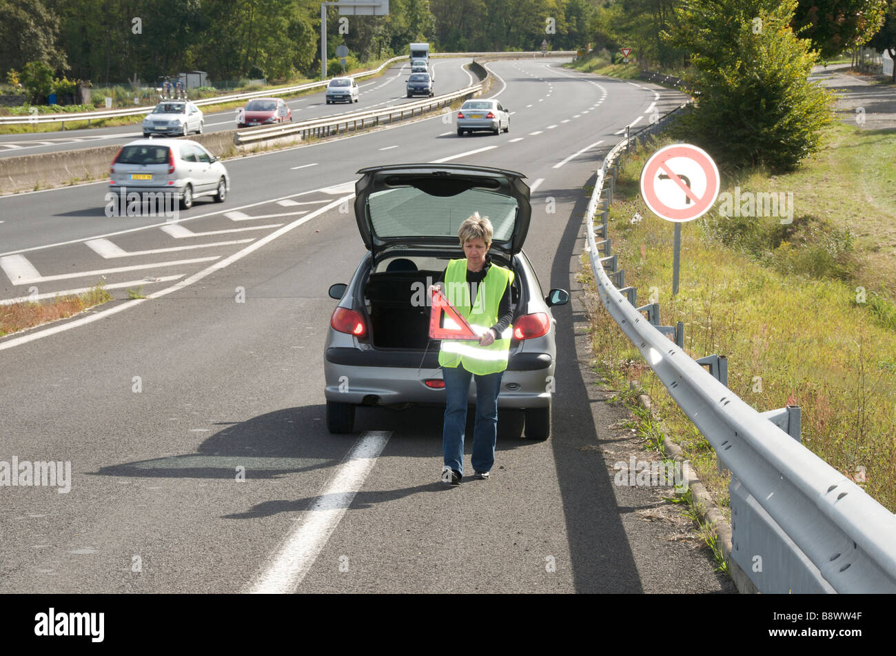 Panne - Frau, die ihr rotes Warndreieck hinter ihrem kaputten Auto auf einer Zufahrtsstraße auf eine französische Autoroute platziert Stockfoto